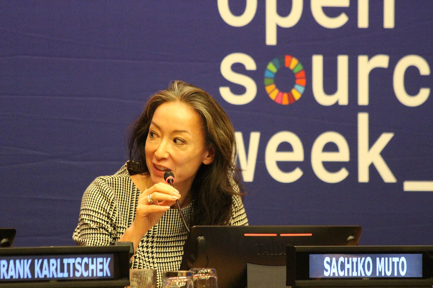 A woman speaks into a microphone at a conference table, with a blue open source week banner behind her and nameplate reading SACHIKO MUTO in front.