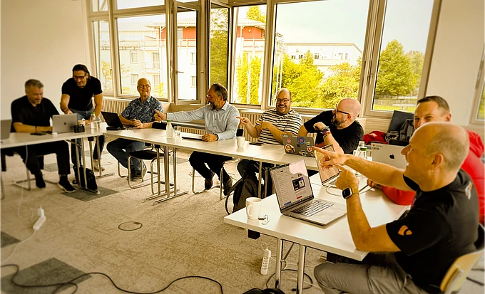 Seven men sit around a U-shaped table in a bright meeting room, engaging in discussion and laughter, with laptops and coffee cups in front of them. Large windows show a view of trees and buildings outside.