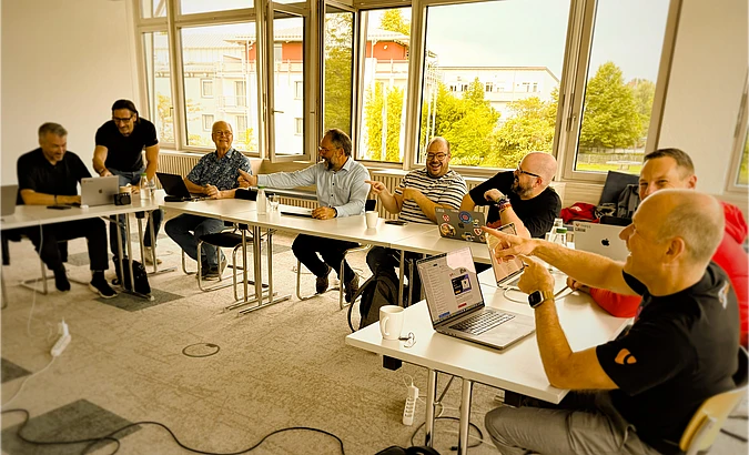 Seven men sit around a U-shaped table in a bright meeting room, engaging in discussion and laughter, with laptops and coffee cups in front of them. Large windows show a view of trees and buildings outside.