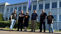 Six people stand outside in front of a modern building with large windows and a flag. They are casually dressed and smiling on a sunny day, with greenery and blue sky in the background.