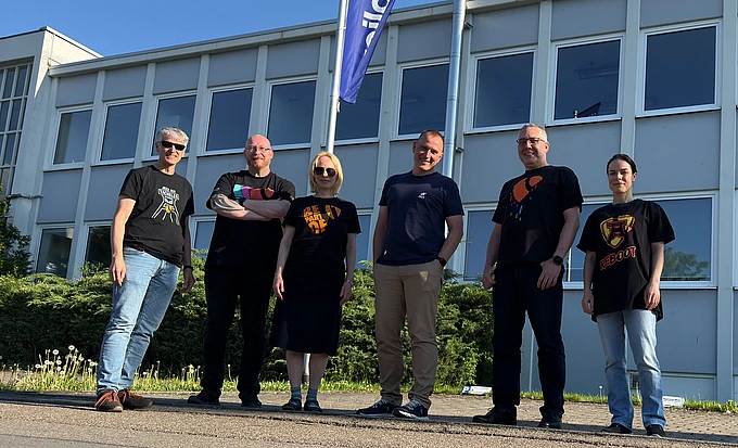 Six people stand outside in front of a modern building with large windows and a flag. They are casually dressed and smiling on a sunny day, with greenery and blue sky in the background.