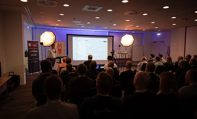 A seated audience faces a speaker and a projection screen in a conference hall, with Contao Summit banners at the side of the stage.