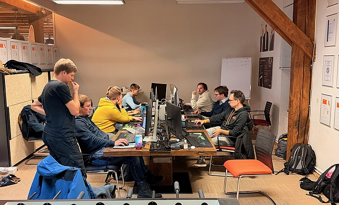 Several people work on computers at a large table in an office. Some are seated, focused on their monitors, while one person stands nearby. Coats, backpacks, and documents are visible in the casual, collaborative workspace.