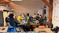 Several people work on computers at a large table in an office. Some are seated, focused on their monitors, while one person stands nearby. Coats, backpacks, and documents are visible in the casual, collaborative workspace.