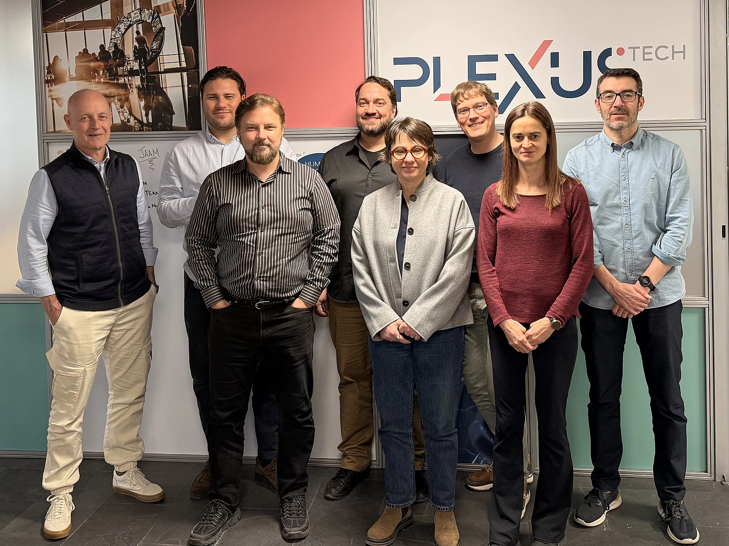 Eight people stand in two rows, smiling, in front of a Plexus Tech sign indoors. They wear casual or business-casual clothing, with neutral and warm colors in an office setting.