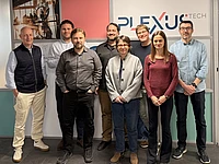 Eight people stand in two rows, smiling, in front of a Plexus Tech sign indoors. They wear casual or business-casual clothing, with neutral and warm colors in an office setting.