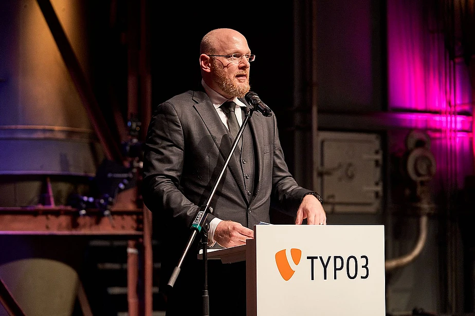 A man in a suit speaks at a TYPO3 podium on a stage.