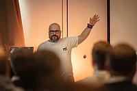Martin Helmich, wearing a white T-shirt, gestures toward the screen while presenting to an audience.