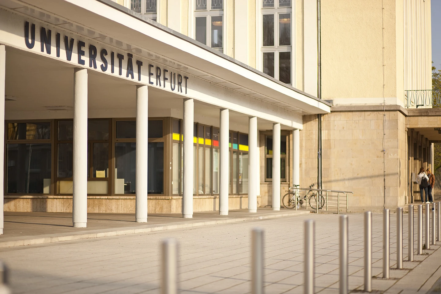 The entrance of Universität Erfurt with white columns, large glass windows, and bicycles parked nearby. Two people stand near the building in the background.