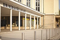 The entrance of Universität Erfurt with white columns, large glass windows, and bicycles parked nearby. Two people stand near the building in the background.