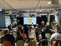A group of people sit facing a projector screen in a modern, well-lit room, watching a presentation titled “TYPO3 Camp.” Two speakers stand at the front near the screen.