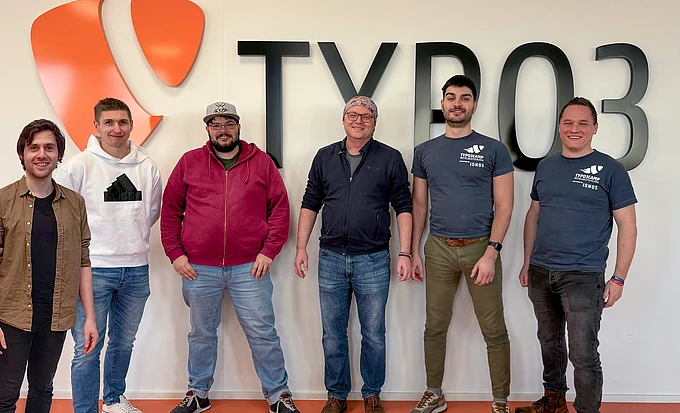 Six people stand smiling in front of a wall with the TYPO3 logo and text. Four of them are in casual clothing, while two wear matching TYPO3 t-shirts.
