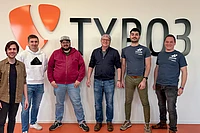Six people stand smiling in front of a wall with the TYPO3 logo and text. Four of them are in casual clothing, while two wear matching TYPO3 t-shirts.