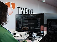A person is working at a computer with code and terminal windows open on the screen, sitting in a modern office space with a large TYPO3 logo on the wall in the background. code sprint community developer laptop typo3 code
