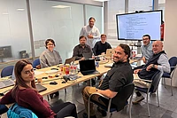 Eight people sit around a conference table with laptops, papers, and drinks, smiling at the camera. A man stands at the end of the table. A large monitor behind him displays a document with text.
