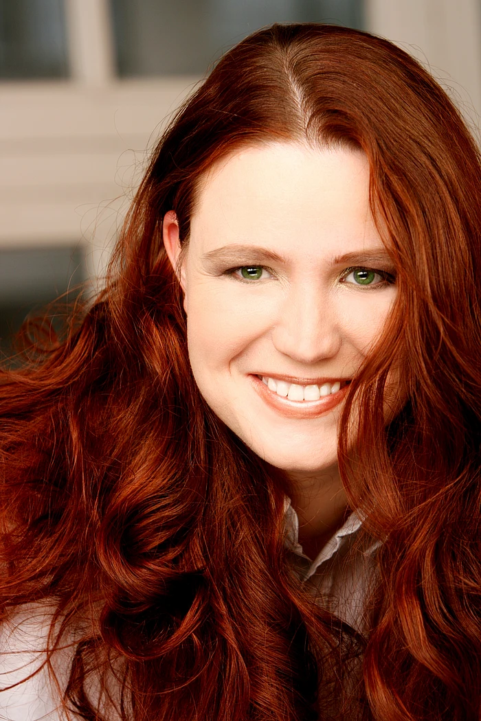 A woman with long, wavy red hair and green eyes smiles brightly at the camera. She is wearing a light-colored shirt and is photographed indoors with a blurred background.