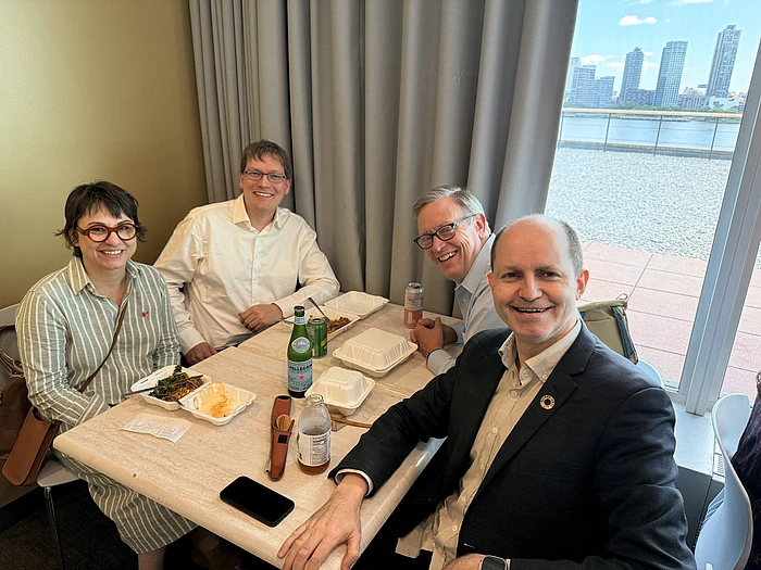 Four people smiling at a dining table with food and drinks, city skyline visible through window behind them