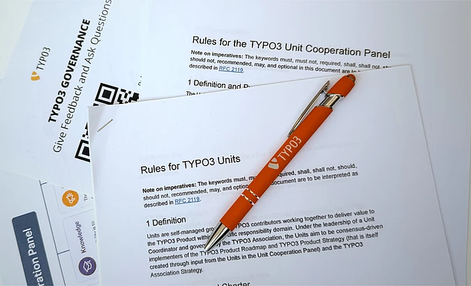 A silver and orange pen labeled TYPO3 rests on printed documents about TYPO3 governance and unit cooperation rules, with a QR code visible on one page.