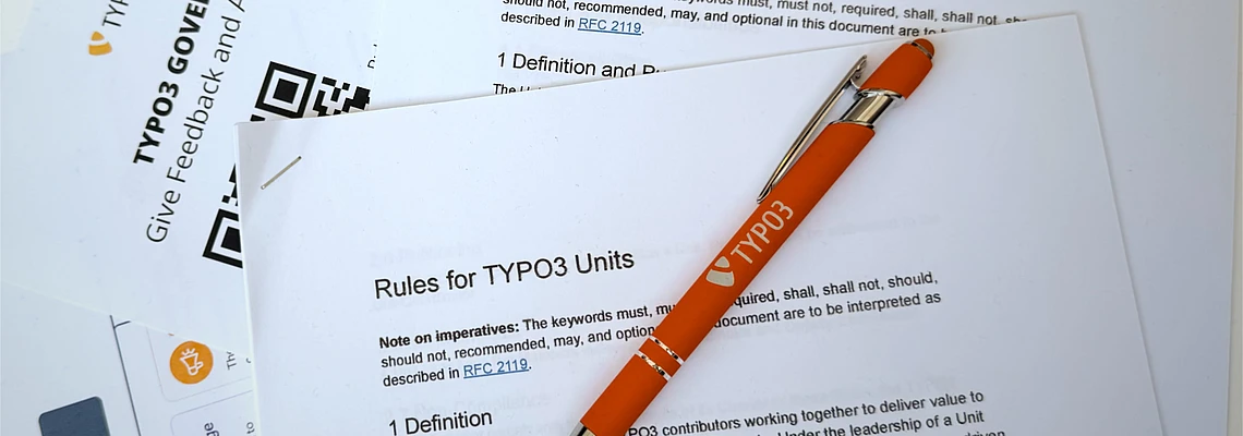A silver and orange pen labeled TYPO3 rests on printed documents about TYPO3 governance and unit cooperation rules, with a QR code visible on one page.