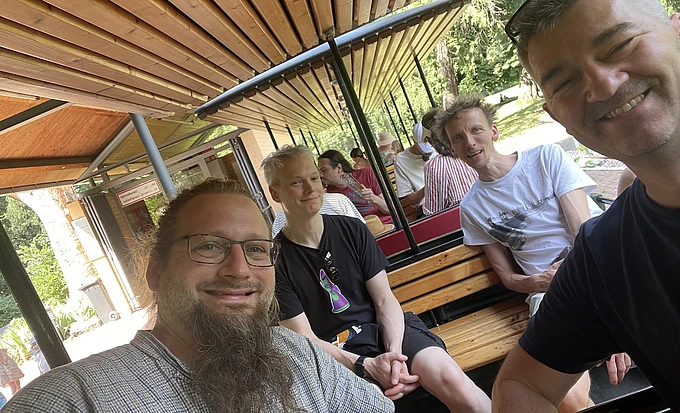 Four people are smiling and sitting on wooden benches inside an outdoor tram or train, with greenery and other passengers visible in the background. The photo is taken in bright daylight.