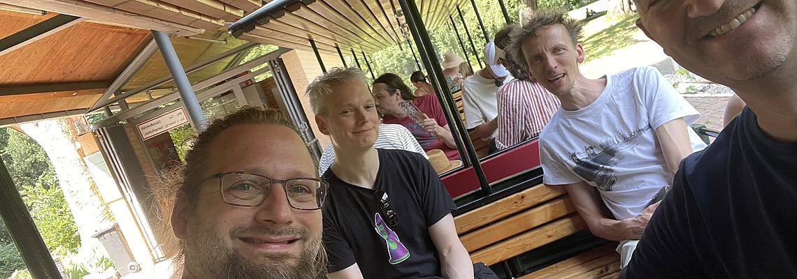 Four people are smiling and sitting on wooden benches inside an outdoor tram or train, with greenery and other passengers visible in the background. The photo is taken in bright daylight.