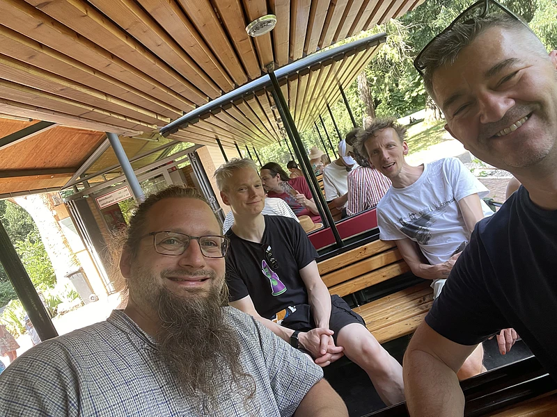 Four people are smiling and sitting on wooden benches inside an outdoor tram or train, with greenery and other passengers visible in the background. The photo is taken in bright daylight.