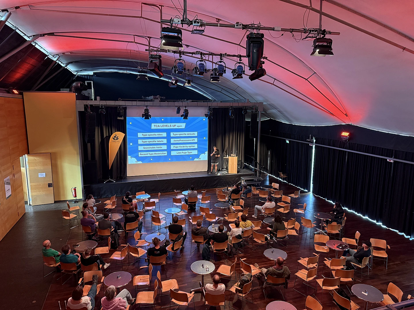 Conference presentation on TCA Levels Up with audience seated in orange chairs under pink-lit ceiling with stage and projection screen.