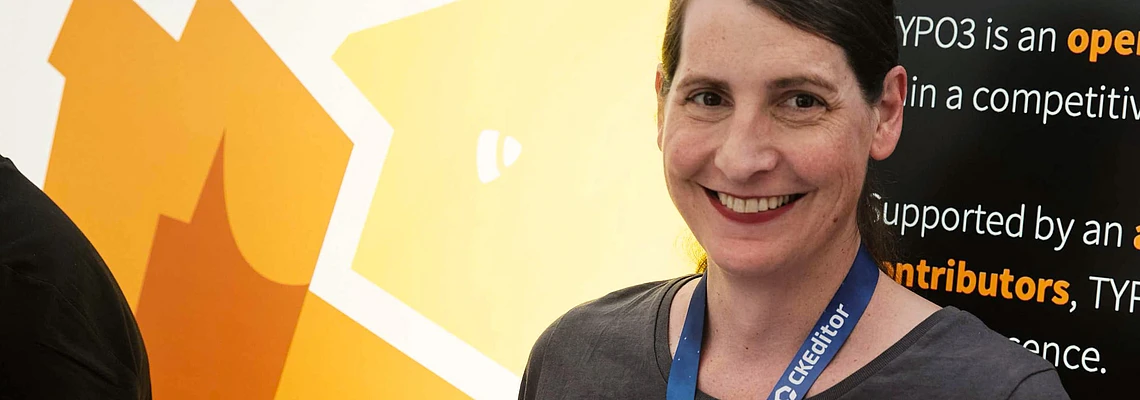 Woman wearing TYPO3 conference badge and t-shirt smiling at camera with yellow TYPO3 logo and informational banner in background