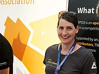 Woman wearing TYPO3 conference badge and t-shirt smiling at camera with yellow TYPO3 logo and informational banner in background