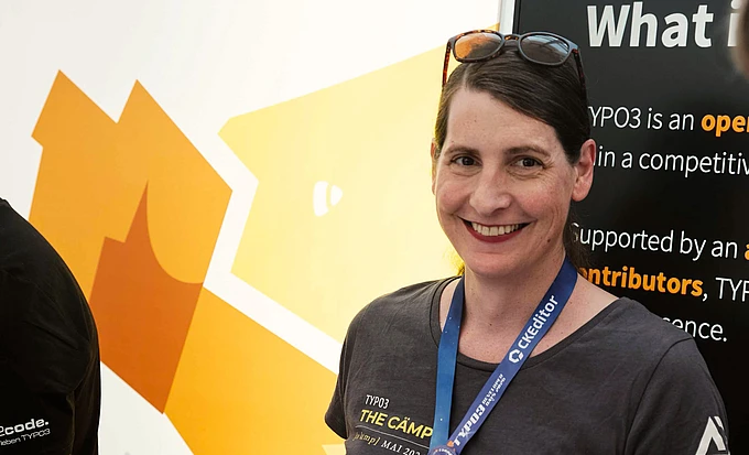 Woman wearing TYPO3 conference badge and t-shirt smiling at camera with yellow TYPO3 logo and informational banner in background