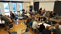 A group of people are seated in small groups around tables in a conference room, engaged in discussion. The atmosphere appears collaborative and professional.
