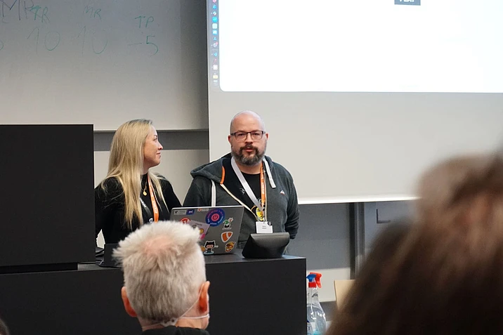 Two people stand at a podium presenting to an audience at a conference. One is speaking while the other listens. A laptop with stickers is on the podium, and a projector screen is in the background.
