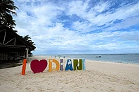 Colorful 'I love Diani' sign on sandy beach with palm trees, ocean, and blue sky
