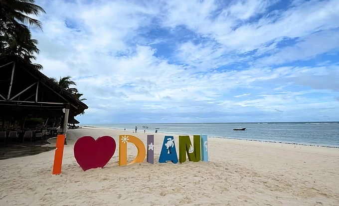 Colorful 'I love Diani' sign on sandy beach with palm trees, ocean, and blue sky