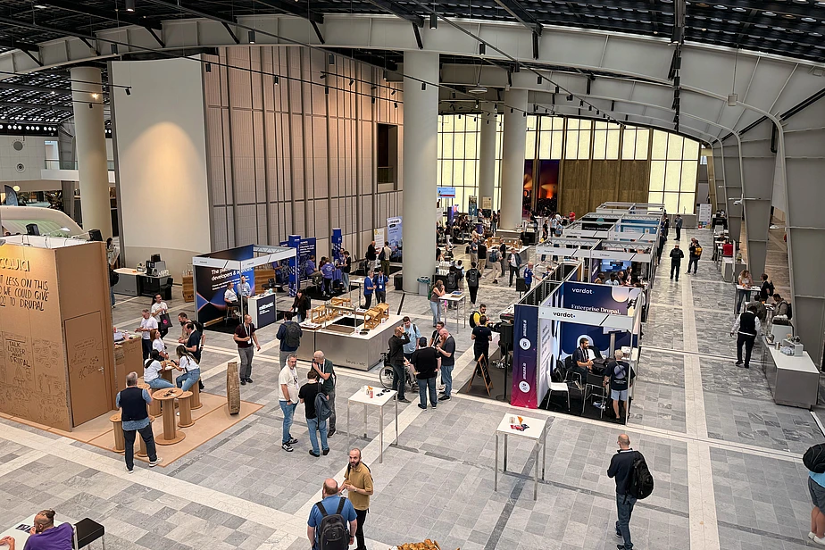 Overhead view of a large indoor trade show or conference with multiple exhibition booths, attendees walking around, and modern industrial architecture with skylights.