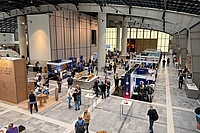 Overhead view of a large indoor trade show or conference with multiple exhibition booths, attendees walking around, and modern industrial architecture with skylights.