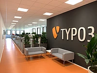 Bright office hallway with orange flooring, gray seating, plants, and a large TYPO3 sign on a dark wall.