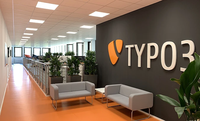 Bright office hallway with orange flooring, gray seating, plants, and a large TYPO3 sign on a dark wall.