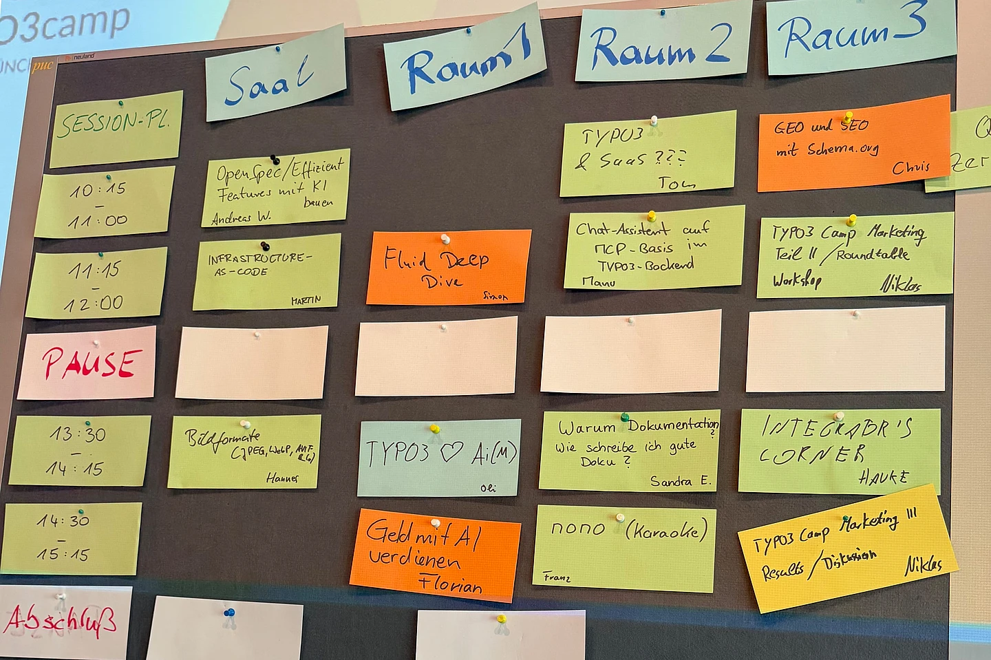 Conference schedule board with sticky notes organizing sessions across multiple rooms (Saal, Raum 1-3) with times, topics, and speaker names.