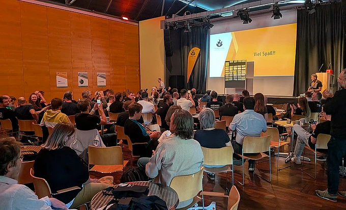 Large audience seated at TYPO3 camp munich in modern auditorium with orange walls watching presentation on yellow screen displaying 'Viel Spaß!!'