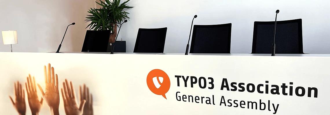 A conference panel table with four chairs and microphones, labeled TYPO3 Association General Assembly. The front features images of raised hands. A potted plant and lamp are in the background.