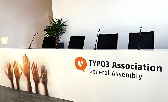 A conference panel table with four chairs and microphones, labeled TYPO3 Association General Assembly. The front features images of raised hands. A potted plant and lamp are in the background.