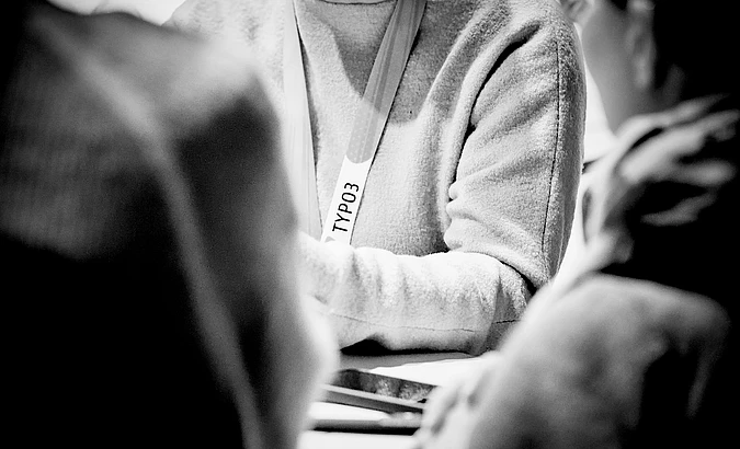 Person wearing TYPO3 lanyard in black and white photo, surrounded by others in casual setting