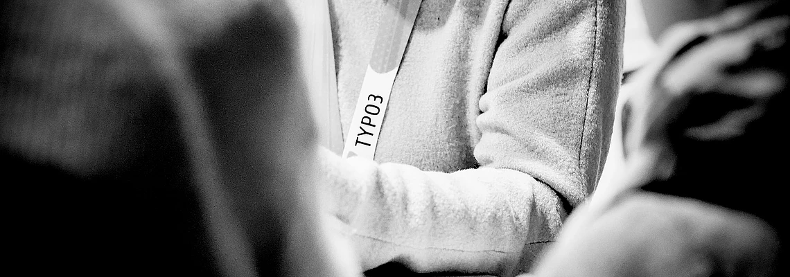 Person wearing TYPO3 lanyard in black and white photo, surrounded by others in casual setting
