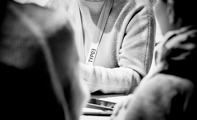 Person wearing TYPO3 lanyard in black and white photo, surrounded by others in casual setting