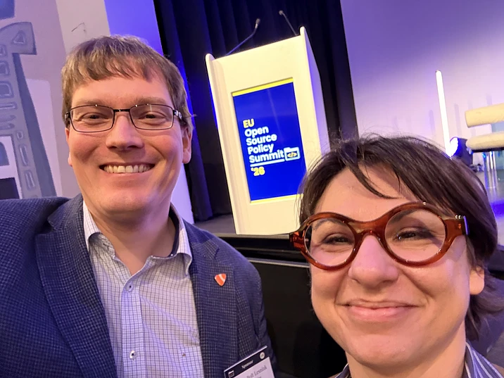 Two people smiling at EU Open Source Policy Summit 2023 event with blue and yellow branding backdrop