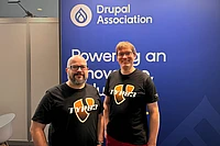 Two men wearing TYPO3 t-shirts stand in front of a Drupal Association banner at a conference