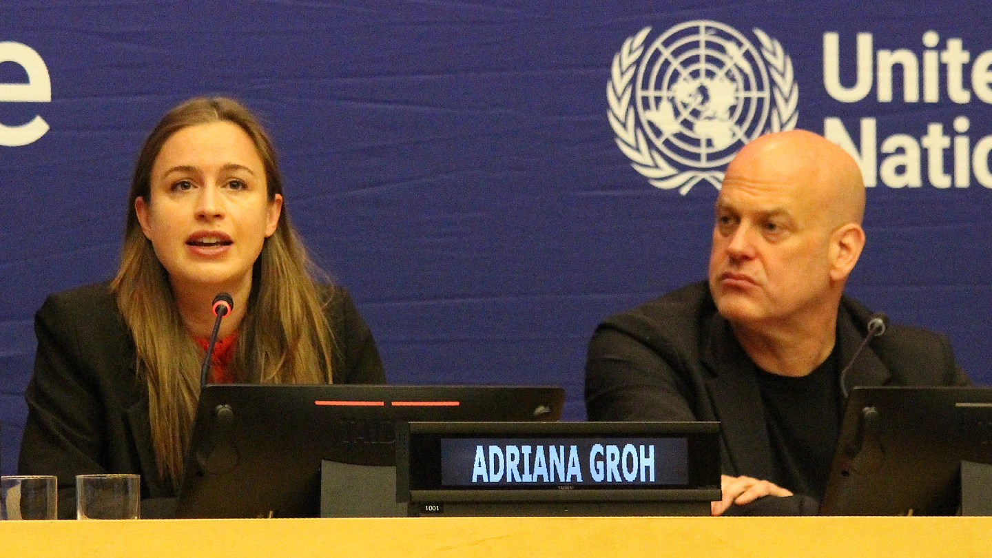 A woman speaks into a microphone at a UN conference panel, while a man sits beside her. A nameplate reads Adriana Groh. The United Nations logo is visible on the blue backdrop behind them.