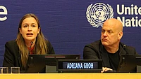 A woman speaks into a microphone at a UN conference panel, while a man sits beside her. A nameplate reads Adriana Groh. The United Nations logo is visible on the blue backdrop behind them.